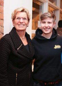 Kathleen Wynne and Stella Green pose for the camera, smiling. Both are wearing black and have short blonde hair. Kathleen is wearing red rimmed glasses.
