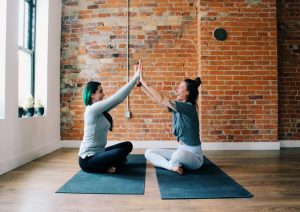 Two people sitting on yoga mats facing each other with their hands raised and pals together in front of them.