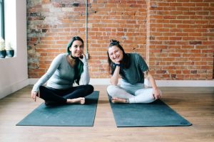 Two people sitting cross legged on black yoga mats smiling into the camera. Behind them is a red brick wall.