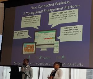 Tendai & Funmi up on a stage presenting with a large projection screen behind them that reads "NexJ Connected Wellness: A Young Adult Engagement Platform"