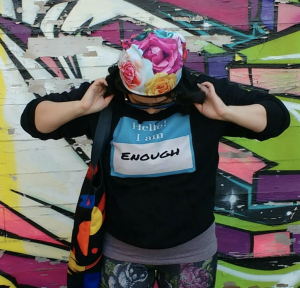 A photo of Amanda Lederle, wearing a floral cap, looking down at their shirt which reads "Hello! I am enough"