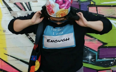 A photo of Amanda Lederle, wearing a floral cap, looking down at their shirt which reads "Hello! I am enough"