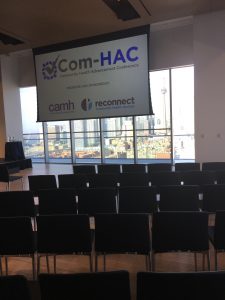 An empty room setup for an event with chairs in the foreground and a large projector screen hung from the ceiling at the front that reads "Com-HAC"