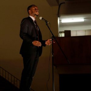 Funmi, dressed in a black suit, speaking into a microphone