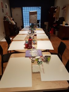 A photo of a n empty room, looking down two tables prepared with blank sheets of paper set out and groups of art supplies.