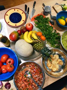 A top down photo of a table full of food. There's a fruit bowl with bananas and pineapple, a bowl with dip and chips, a platter of greens and a bowl of tomatoes.