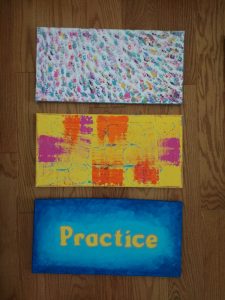 3 rectangular canvases stacked on top of each other on a wooden surface. The top one is a bunch of small textured stokes in different colours. The middle is yellow canvas with brushstrokes of pink and orange. The bottom is a blue painted canvas with yellow text in the middle that reads "Practice"