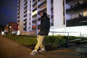 Jah-Reign sitting and leaning against a handrail. In the background is a condo building, lit up by lights in the night.