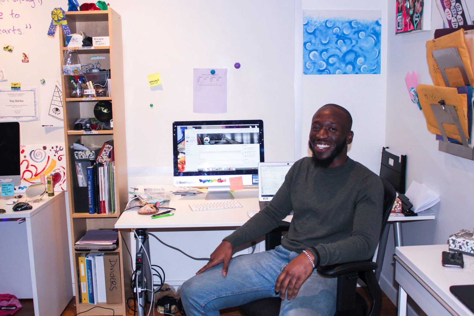 A portrait of staff member Funmi, who is sitting down at a desk with a computer, with lots of knick knacks, artwork and papers covering office walls.