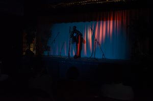 A dark photo showing a person standing up on stage in front of a microphone.