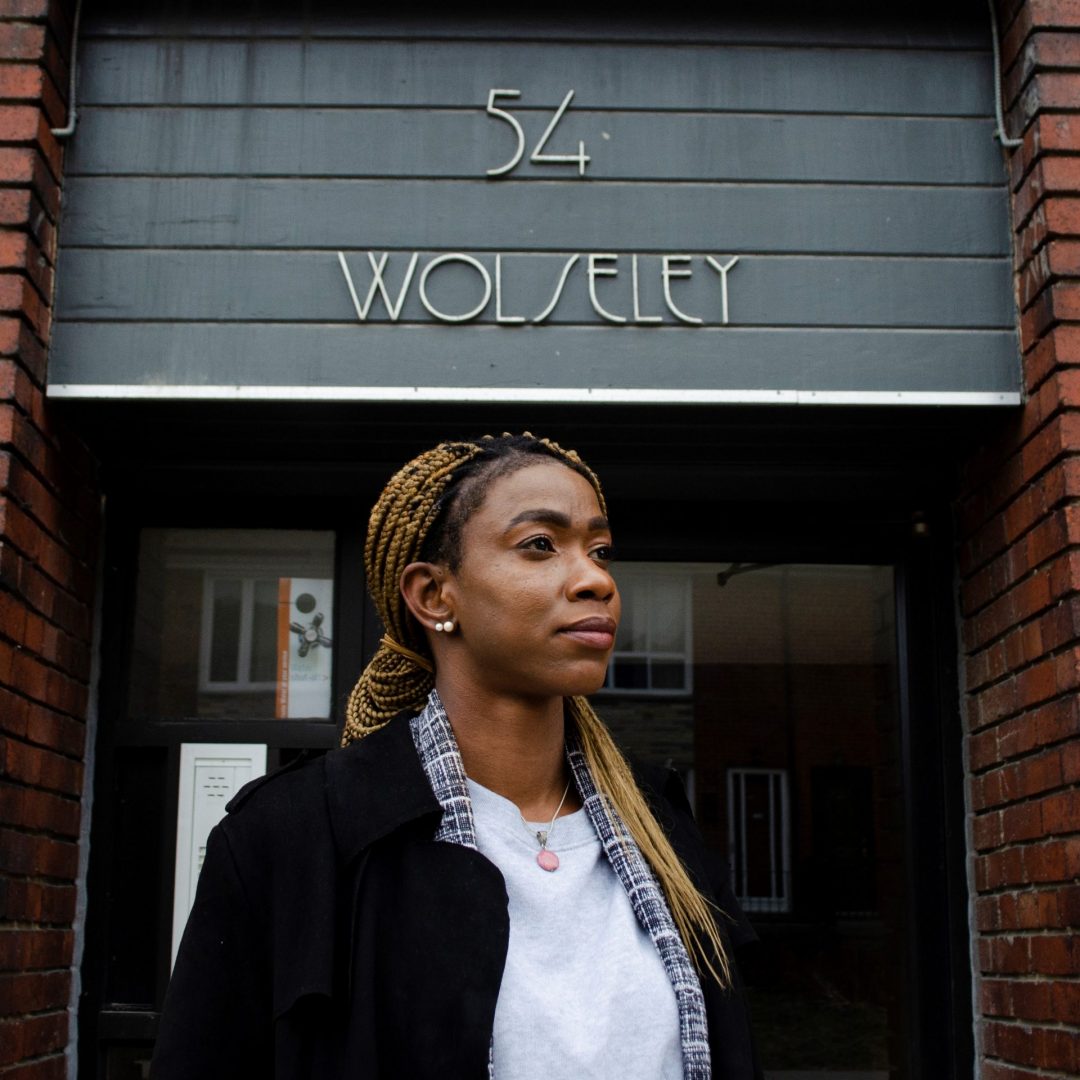 Portrait of Lereen standing outside of 54 Wolseley Street, the future home of Stella's Place. Lereen is looking to the right off into the distance with a serious, determined expression on her face. She has two pearl earrings in her ears and long hair hanging over one shoulder, wearing a white shirt with a black coat overtop. Behind her is the building with 54 Wolseley written above her.