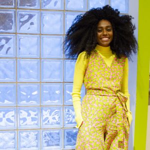 A portrait of Jennille standing to the right of the photo, wearing a bright lime green outfit and standing in front of a wall of square tiled windows.