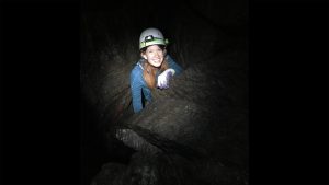 Photograph of Carol Krause in a dark cave. She is fair skinned with long orange hair, in this photo she is wearing a light blue sweater and has a big smile on her face, showing all of her teeth. On her head is a safety helmet with a light attached that is the only source of light in the photo. She has white gloves on her hand and is holding onto the rock in front of her. Only the top half (above her torso) is showing and the rest is behind more rocks.