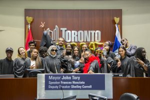 A group photo of the Community Healing Project team, all making funny faces for the camera, and dressed in black graduation gowns and some wearing feather boas and sunglasses. they are standing in front of a city of Toronto sign and behind a podium that says "Mayor John Tory"