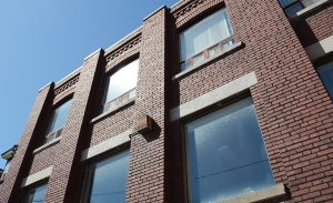 A photo taken from the outside of the new Stella's Place building. It's taken from right outside the building looking up, giving it a feeling of growth. The brick is red and the sky is bright blue which is reflected onto the windows.