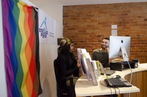 Two Stella's Place Access Coordinators sitting behind the front desk in Stella's Place front cafe space. The Access Coordinators are sitting down speaking to each other. Behind them is a large rainbow Pride Flag, a poster with Stella's Place logo and a red brick wall.