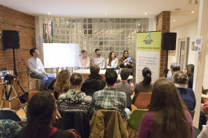 A photo of a panel of speakers in the Stella's Place Café in front of an audience sitting in chairs in the foreground.