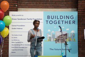 Lereen speaks at the Building Together event, standing in front of a microphone. Behind Lereen is a large banner that reads "Building Together" and to the left, a bouquet of brightly coloured balloons.