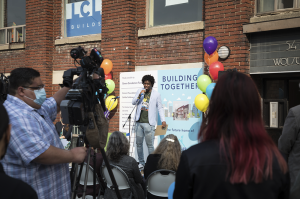 Image of Asante outside the new building at the launch event. Asante is speaking into a microphone on a stage with an audience in the foreground, and a camera crew. You can see the label '54 Wolseley' on the red brick building and some of the windows. Behind Asante is a big graphic board that says 'Building Together' framed by colourful balloons.