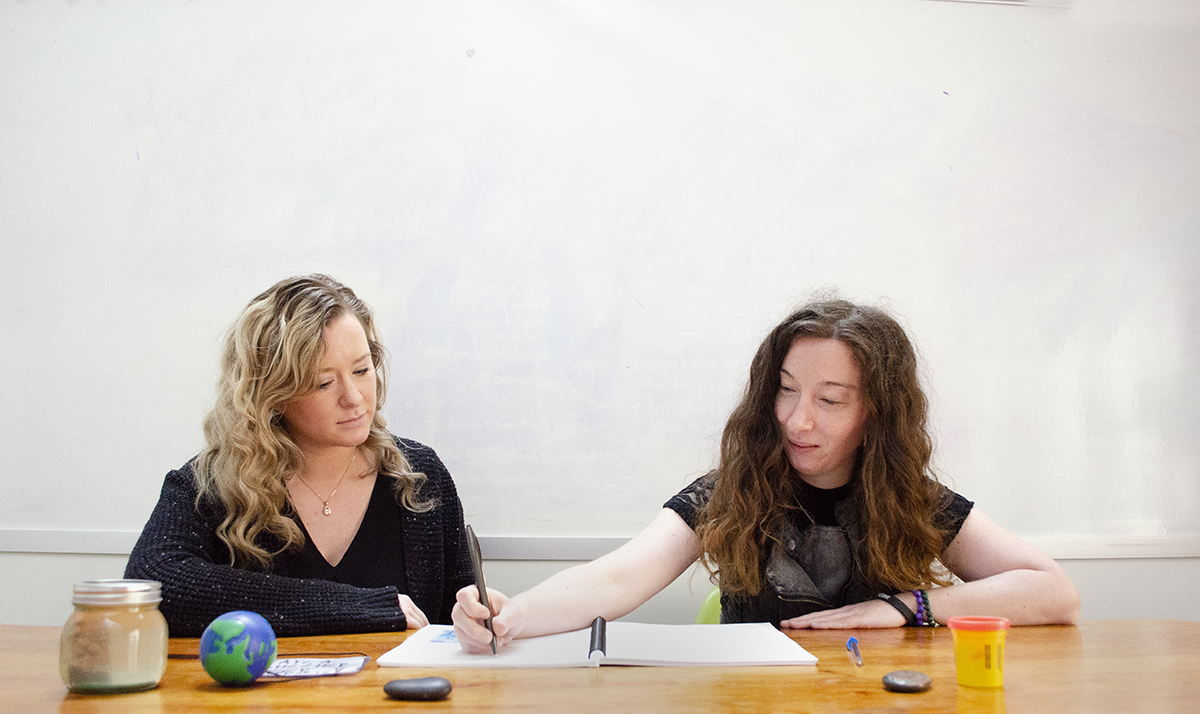 A Stella's Place peer supporter and clinician sitting down together, looking over course material. In front of them on the table are objects used in therapy, such as stress balls, a jar of glitter and rocks.