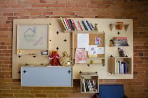 A photo of a wall in the Stella's Place café. A very bright and vibrant wooden shelf hung up on the wall has books, stuffed animals, drawings and more plastered all over.