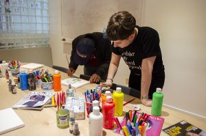 A photo of 2 Stella's Place Peer Support Workers painting, with a big spread of colourful art supplies spread out in front of them.