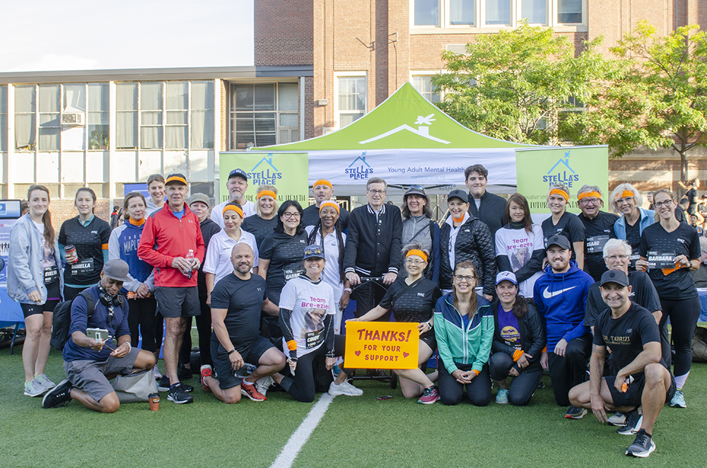 A group photo of a few dozen people smiling into the camera. The photo was taken outside on a sports field outside of a red brick school before a 5K run. John Tory is in the middle of the photo and behind them all is the Stella's Place tent with the logo and two banners on either side.