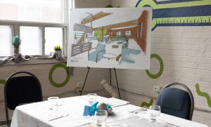 An image from an event at Stella's Place showing an empty prepared table with a tablecloth and table set. Behind the table and beside a window is a big canvas of a sketch of the new Stella's Place building, sitting on an easel.