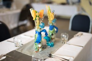 A closeup photo of a table centerpiece. The focus is on the decoration which is a clear vase filled with colourful objects and slices of orange and grapes on sticks standing in it, and tied in a dark blue bow. The table around it is blurred out.