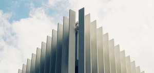 A photo of a modern image against a cloudy blue sky. The image is of the building on an angle, and the walls have dark grey panels in rows.