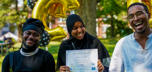 A portrait of 3 people laughing. The person in the middle is wearing a black headscarf and is holding a white paper certificate up. The person on the left is also wearing a head covering and a black shirt with a silver necklace. The person on the right is wearing glasses and a white button up shirt. They are standing in an outside space in summer, the background is filled with green grass and trees.