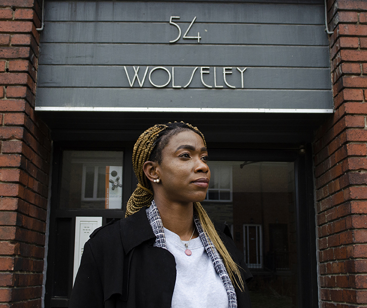 Portrait of Lereen standing outside of 54 Wolseley Street, the future home of Stella's Place. Lereen is looking to the right off into the distance with a serious, determined expression on her face. She has two pearl earrings in her ears and long hair hanging over one shoulder, wearing a white shirt with a black coat overtop. Behind her is the building with 54 Wolseley written above her.Portrait of Lereen standing outside of 54 Wolseley Street, the future home of Stella's Place. Lereen is looking to the right off into the distance with a serious, determined expression on her face. She has two pearl earrings in her ears and long hair hanging over one shoulder, wearing a white shirt with a black coat overtop. Behind her is the building with 54 Wolseley written above her.
