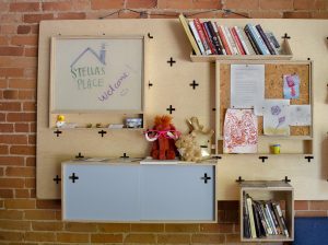 A photo of a wall in the Stella's Place café. A very bright and vibrant wooden shelf hung up on the wall has books, stuffed animals, drawings and more plastered all over.
