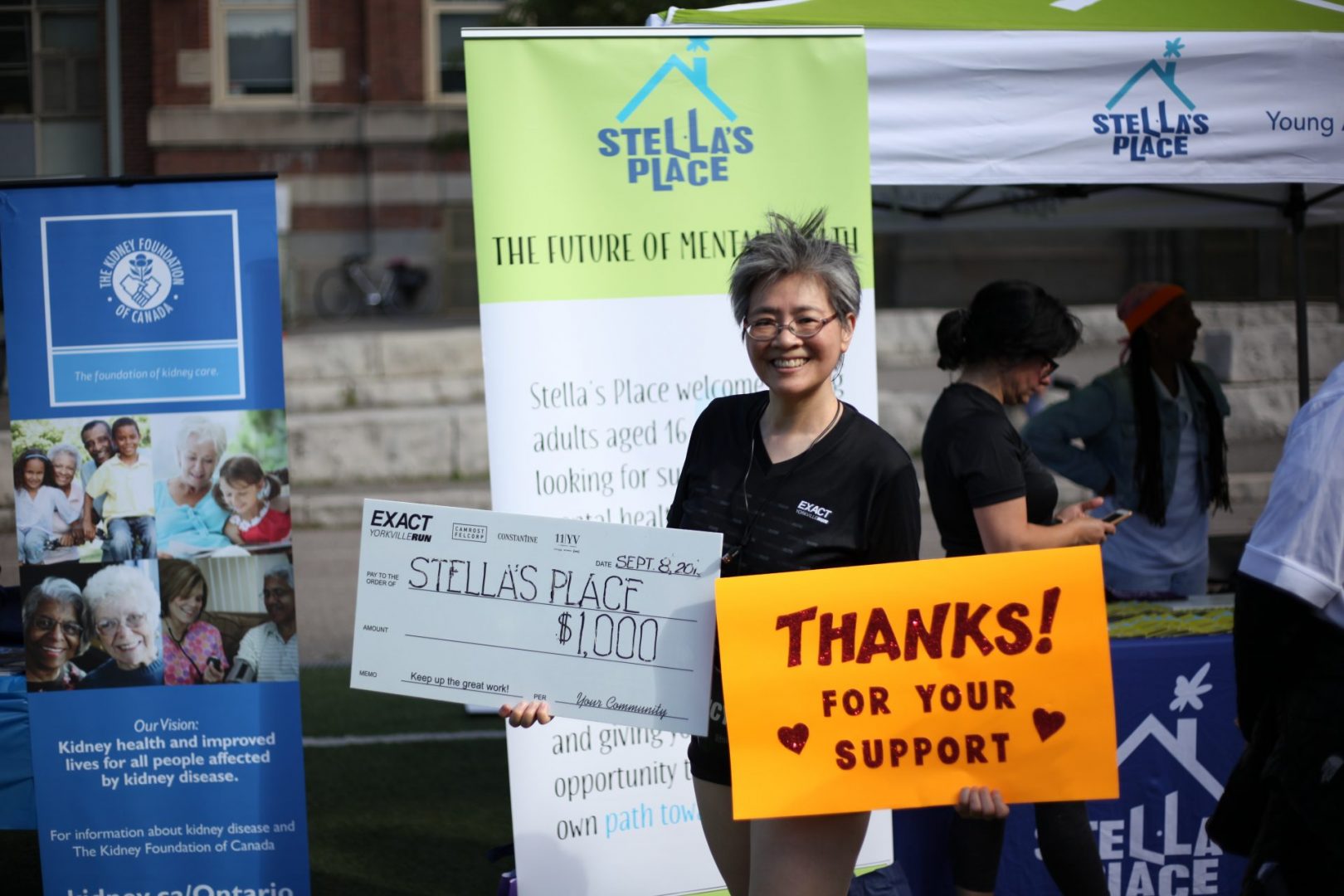 A photo of Stella's Place supporter Nancy Seto holding two signs. The sign on the left is a large cheque poster made out to Stella's Place for $1,000. The poster on the right is an orange sign that says "Thanks for your support" in black.