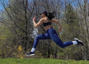 A photo of a young person, mid run, with their arms and legs stretched out in mid air. Behind them is a green forest and you can see the blue sky peeking out from behind the trees. They are wearing blue rights, a cropped black shirt and a ball cap.
