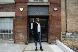 Portrait of Lereen standing outside of 54 Wolseley Street, the future home of Stella's Place. The photo is a full body shot of Lereen, looking to the right off into the distance with a serious, determined expression on her face. Lereen is wearing a black coat and blue jeans and a white shirt underneath. Behind her is the building with 54 Wolseley written above her.