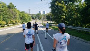 A bunch of people walking on a road on a summer day. The photo is taken from behind with everyone's back facing the camera. Everyone is wearing a white t-shirt with a Stella's Place logo on the back. You can see the open road ahead of them with green trees on the sides and a bright blue sky above.