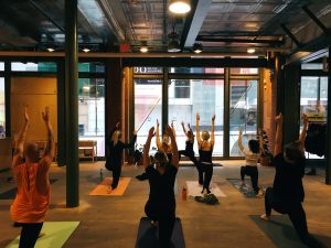 A photo taken of a group of people stretching, it's taken behind the people with their backs facing the camera and looking out through a window. Everyone in the room is on a yoga mat and has their arms stretched out tall above their heads in unison.