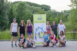 A group of about a dozen people posing for a group photo, standing next to a Stella's Place popup banner. Behind them are green trees in the distance.