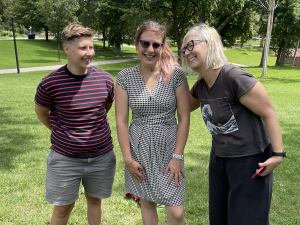 Stella, Corrina and Michelle are standing side by side, talking and laughing, looking at each other. They are standing in a park on a summers day.