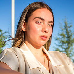 A photo of Mikaila looking into the camera with a serious expression. They are outside with blue sky in the background, and they are wearing a silk white collared shirt and their long brown hair is sitting behind their shoulders.