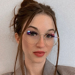 A photo of Samantha with a small smile. Samantha has long brown hair that is pinned up with two strands falling in front of their face, they are wearing clear framed glasses and red lipstick.