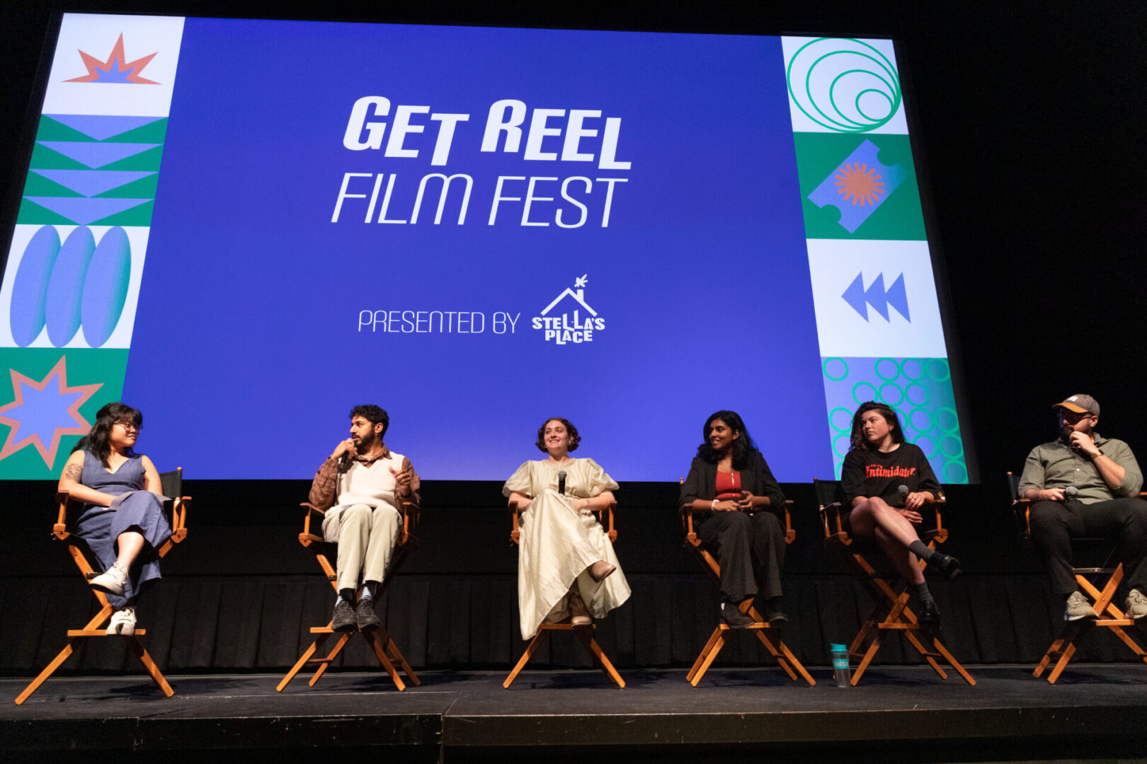 A group of filmmakers sit in director chairs in front of a blue cinema screen that says 'Get Reel Film Fest' brought to you by Stella's Place.